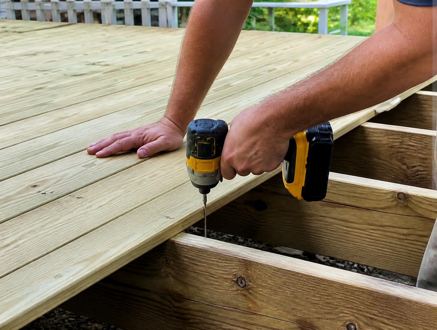 Carpenter fastening a pressure-treated deck board to a joist with a cordless impact driver on a newly framed backyard deck